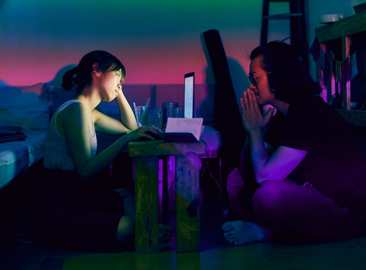 Two people working side by side at a low table in a room washed in neon purple, green and pink light